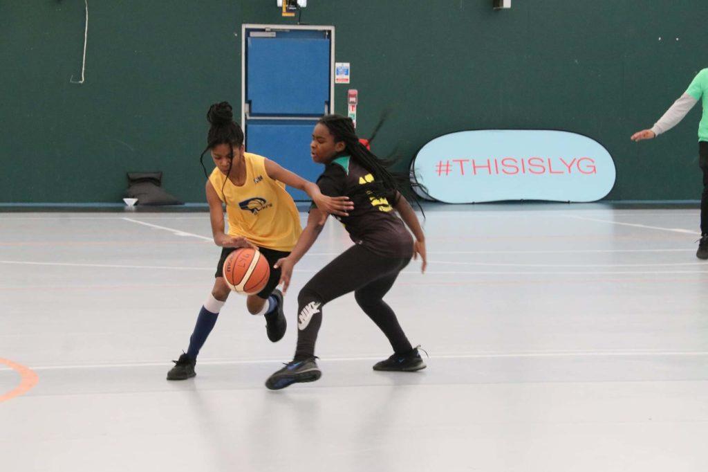 Two girls playing basketball on an indoor court, dribbling and shooting the ball towards the hoop.