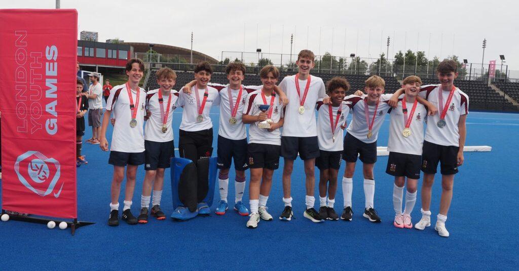 9 white boys and 1 black boy smilae with medals and trophy on hockey pitch